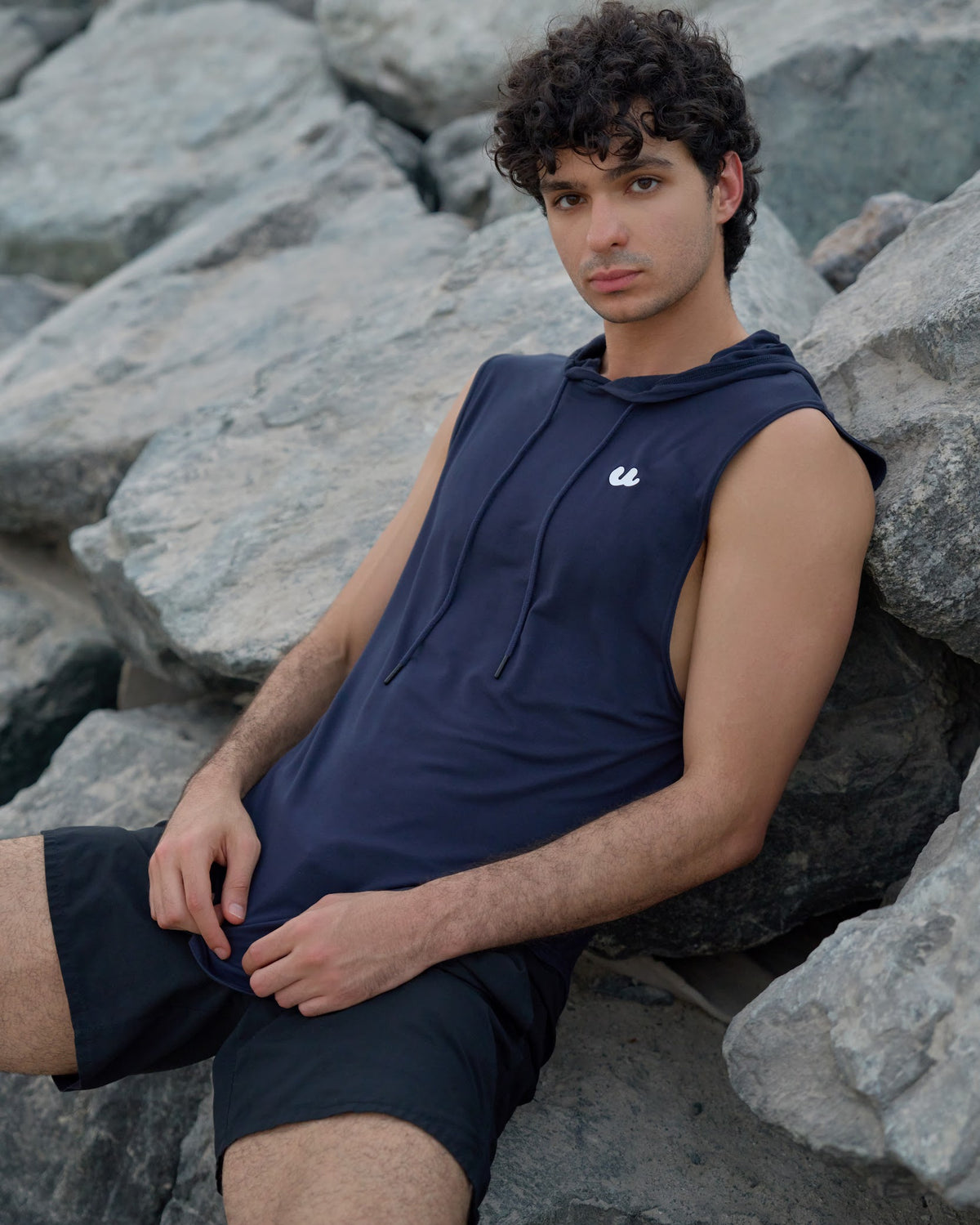 Man in the Urthlete ocean blue sleeveless hoodie resting on rocks by the sea