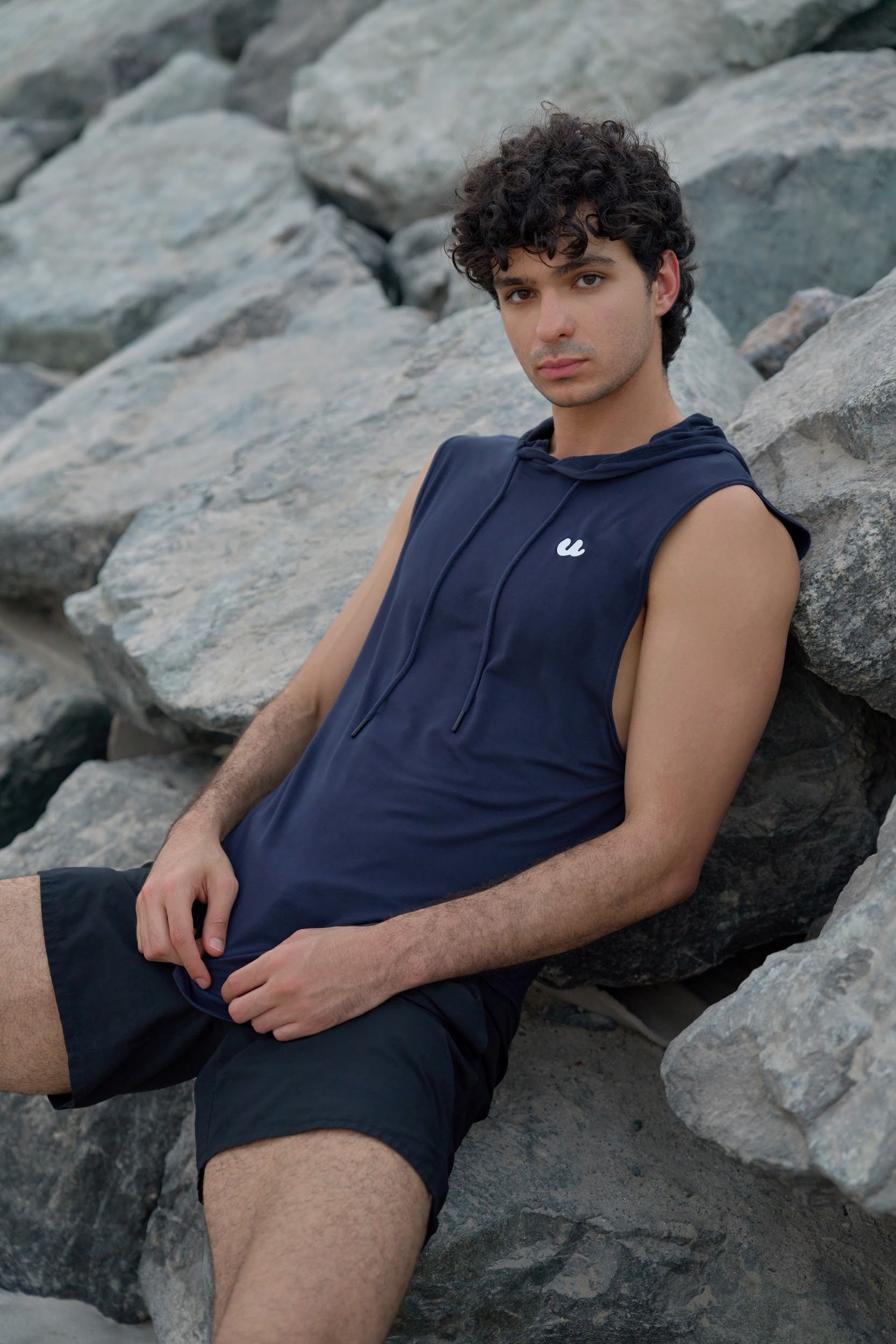 Man in the Urthlete ocean blue sleeveless hoodie resting on rocks by the sea