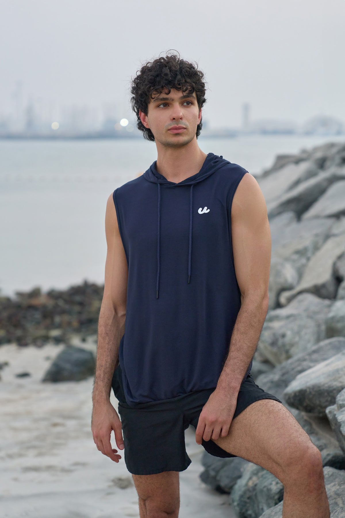 Man in the Urthlete ocean blue sleeveless hoodie standing on rocks by the sea